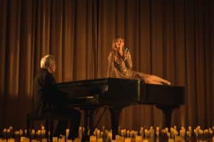 A singer sits on a piano surrounded by candlelight