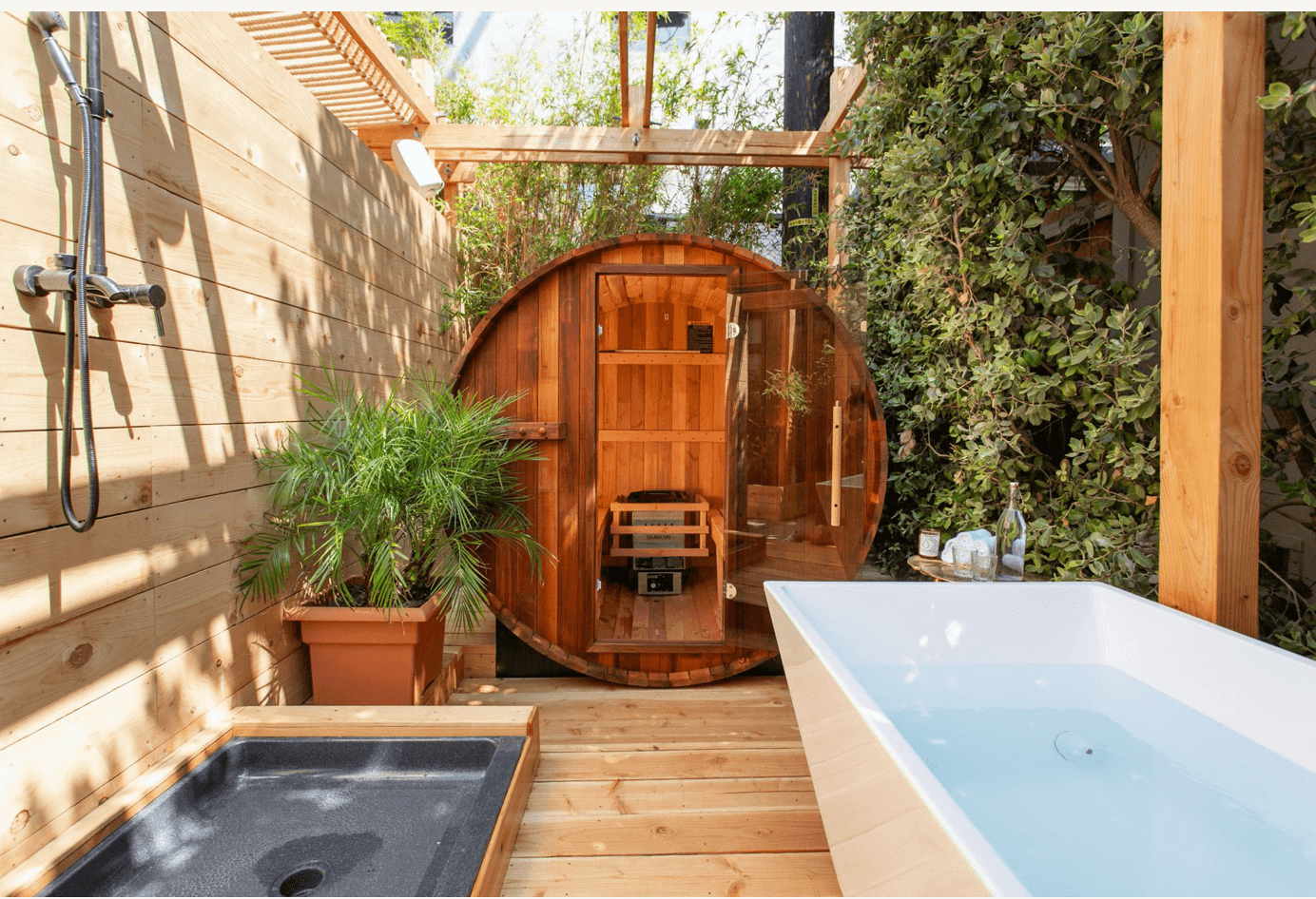 A wooden barrel sauna and outdoor bathtub with greenery