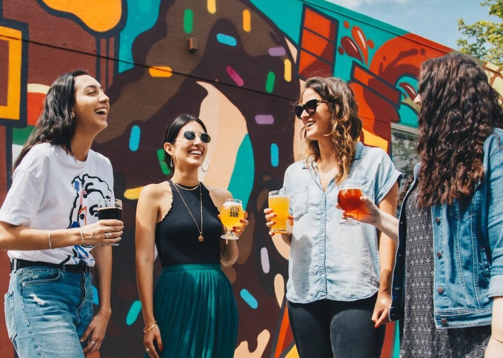 A group of four friends are laughing in front of a colorful mural with beer in their hands.